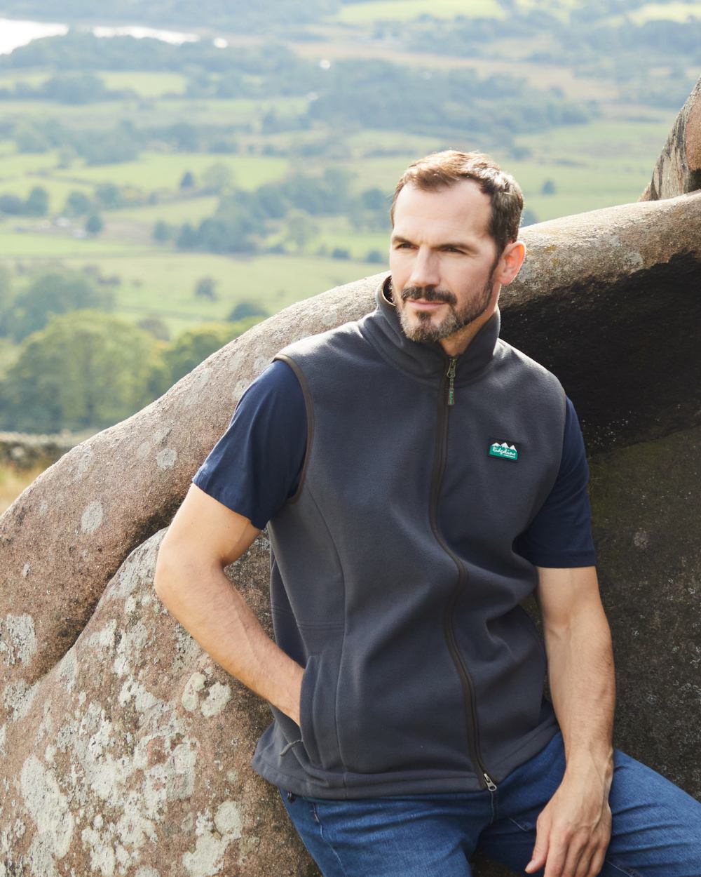 Male model leaning on a rock against a country backdrop wearing a blue ridgeline gilet and jeans.