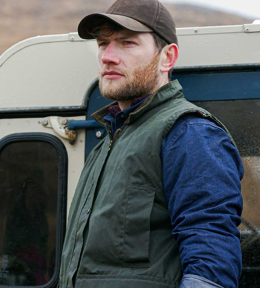 Farmer in Hoggs of Fife waxed cotton gilet stands in front of land Rover Car.