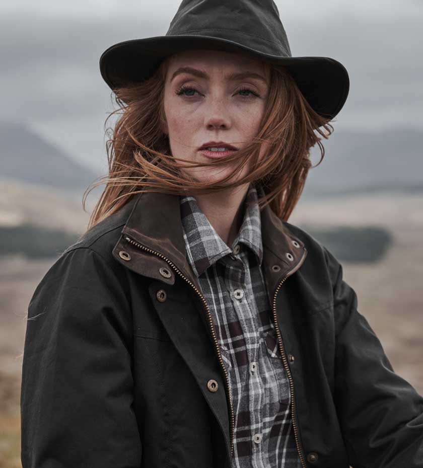 Woman in wax hat and Hoggs waxed jacket over tartan shirt has the wind in her hair and hill in background.