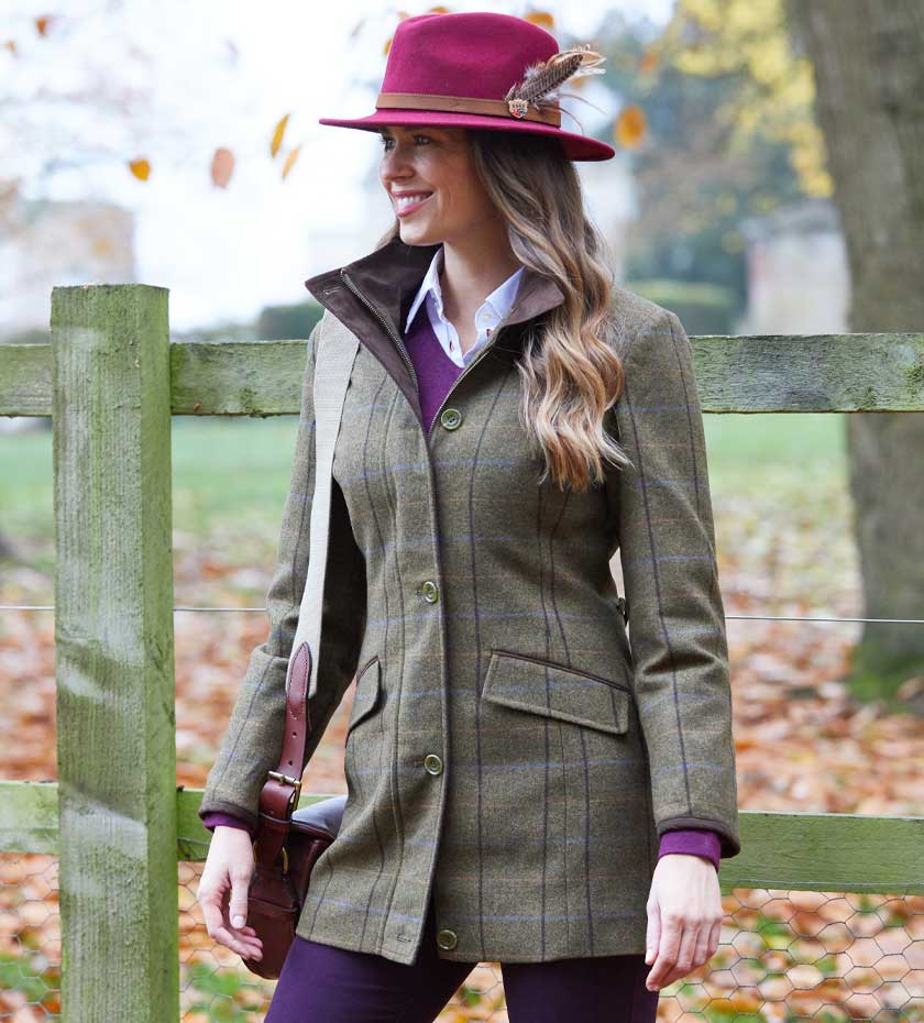 Woman wearing Alan Paine short tweed coat in green and red felt hat with countryside background.