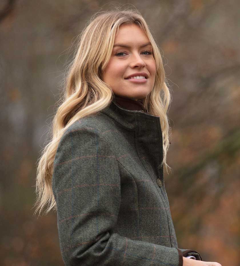 Woman wearing alan Paine combrook tweed coat in green with blurred background.