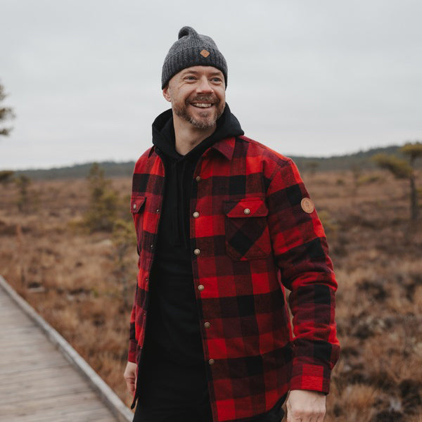 A man wearing a red and black plaid overshirt over a black hoodie and a dark gray knit beanie, standing on a wooden boardwalk in an outdoor marshy landscape.