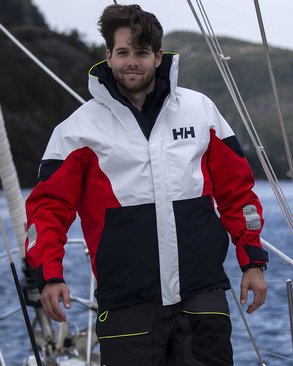 Man wearing Alert Red coloured Helly Hansen Mens Newport Regatta  Jacket on a yacht with sea and headland in the background  