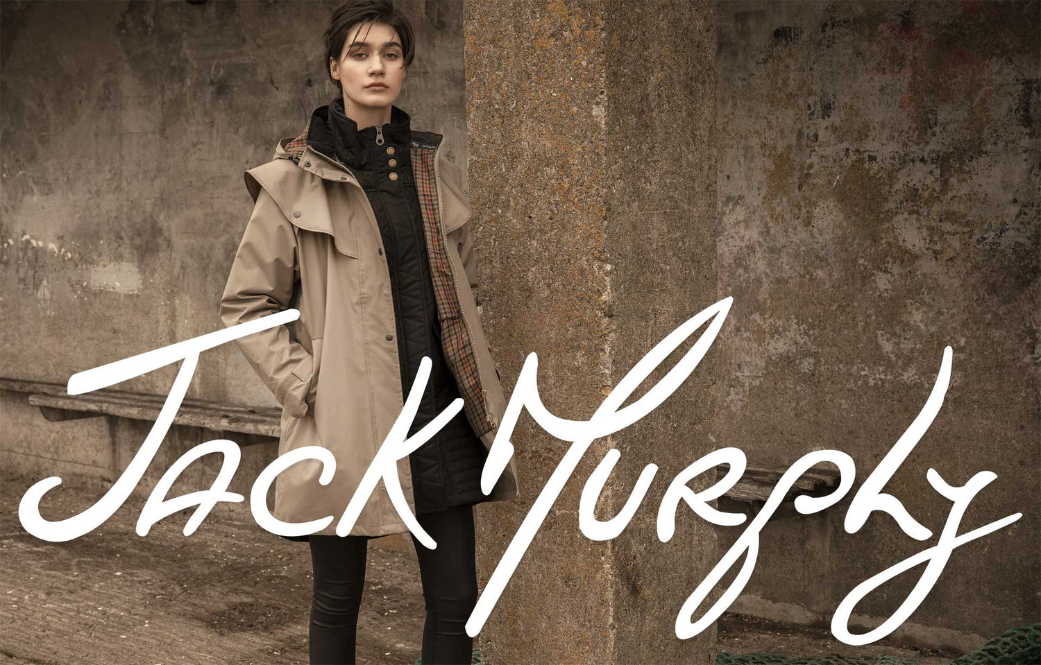 Jack Murphy signature logo in white with woman in beige waterproof coat. Stone wall background.