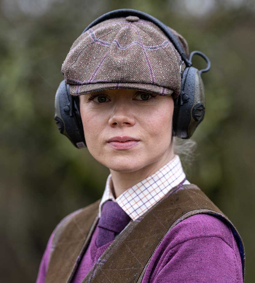 Lady wears a Laksen eight panel tweed country cap and shooting hearing protectors.
