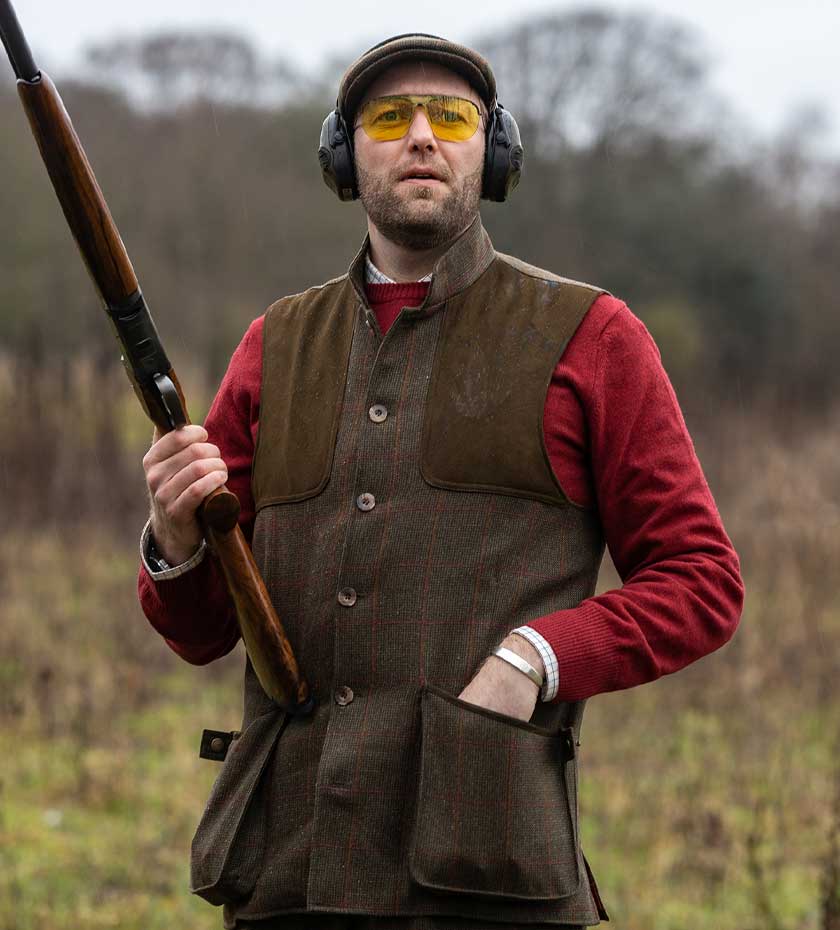Man wearing Laksen Shooting gilet with shoulder pads holding shotgun - mixed woodland background.