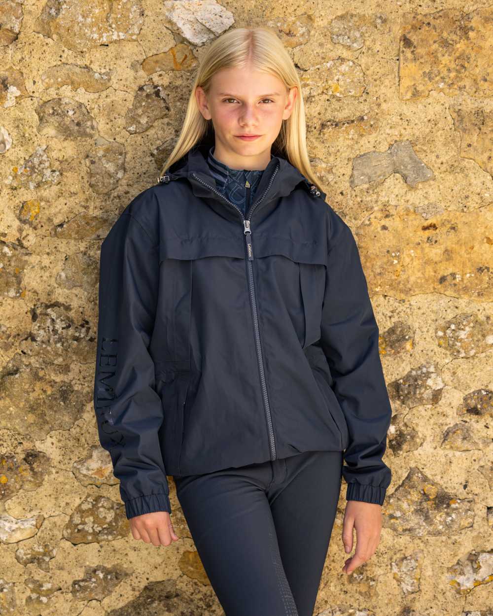 Navy coloured LeMieux Young Rider Dolcie Waterproof Jacket on brick background