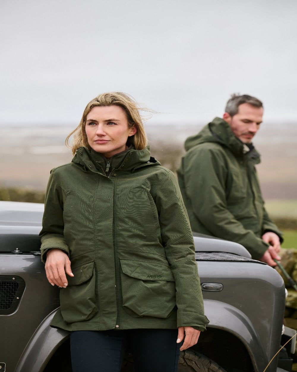 Deep Green Coloured Musto Womens Burnham Waterproof Jacket on Landscape with Vehicle Background