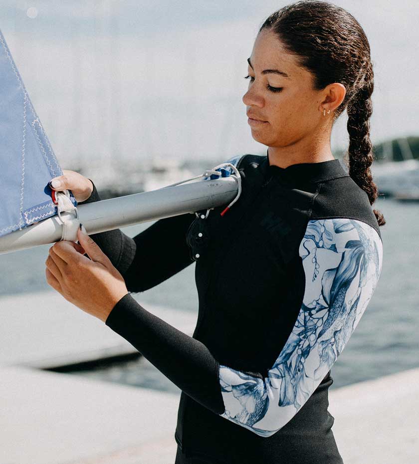 Woman in Black and white sleeved rashguard top adjusts a sail with sea and marina with boats in background.