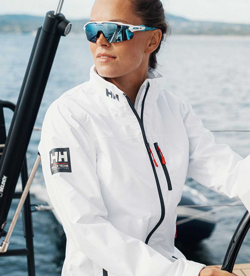 Sailing Mid Layers- Lady in white Helly hansen jacket steers a yacht with sea in background.