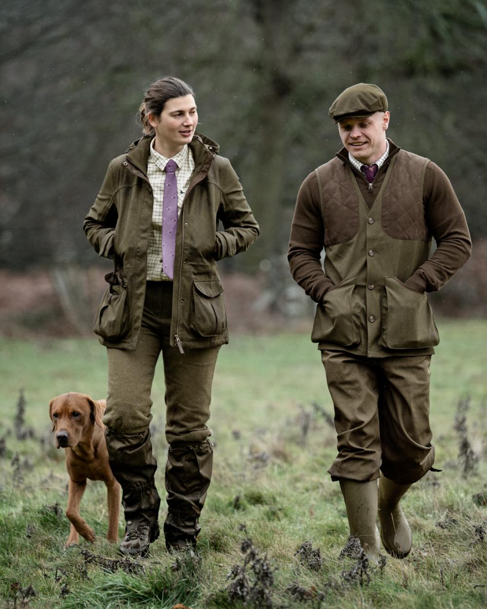 A man and a woman wearing matching brown hunting suits walk in a grassy field with a brown dog. The man wears a flat cap, and both wear olive green jackets, matching trousers, and rubber boots.