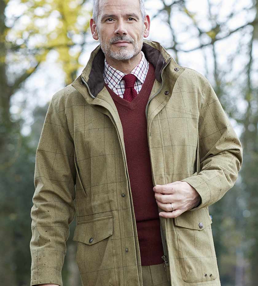 Man wearing Alan Paine jacket with sweater and tattersall shirt with tie, with wood background.