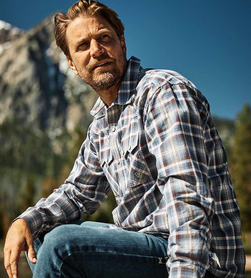Man wears Ariat plaid shirt in grey blue and brown with mountain and sky background.