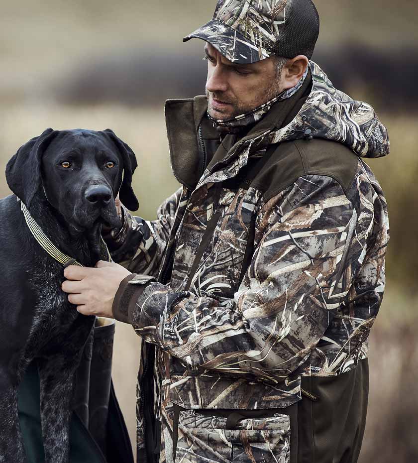 Deerhunter Jackets | man in camouflage hunting jacket pats a black gun dog.