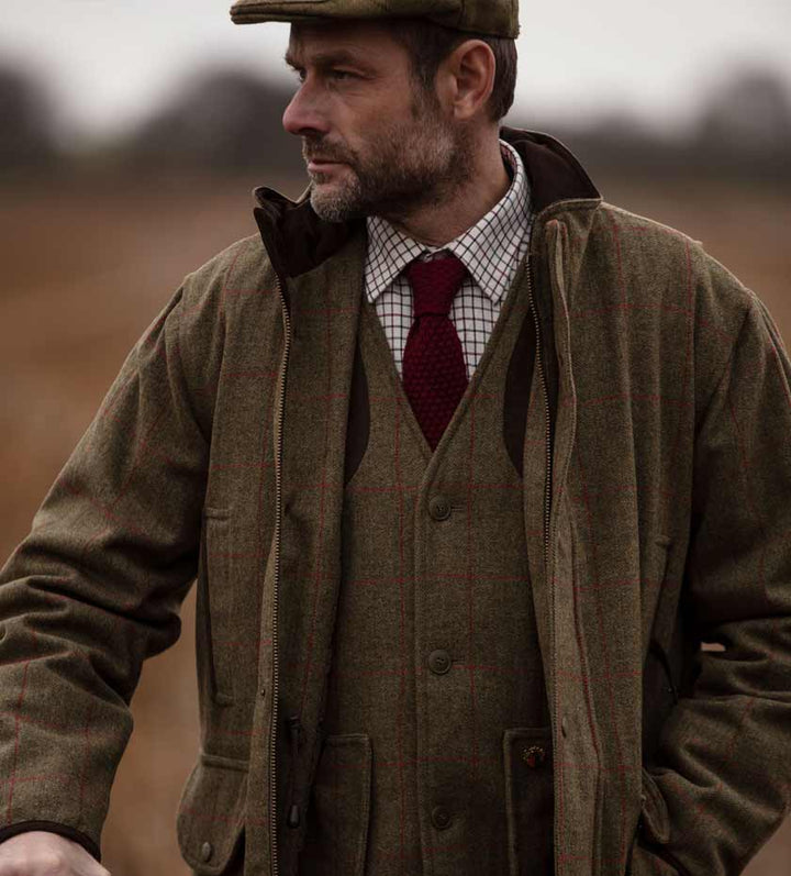 Man wears full tweed outfit of flat cap, waistcoat and jacket over tattersall shirt. Background blurred countryside.