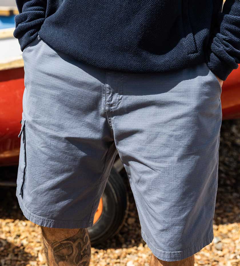Musto blue sailing shorts with cargo pockets on a beach with dinghy boat in background.