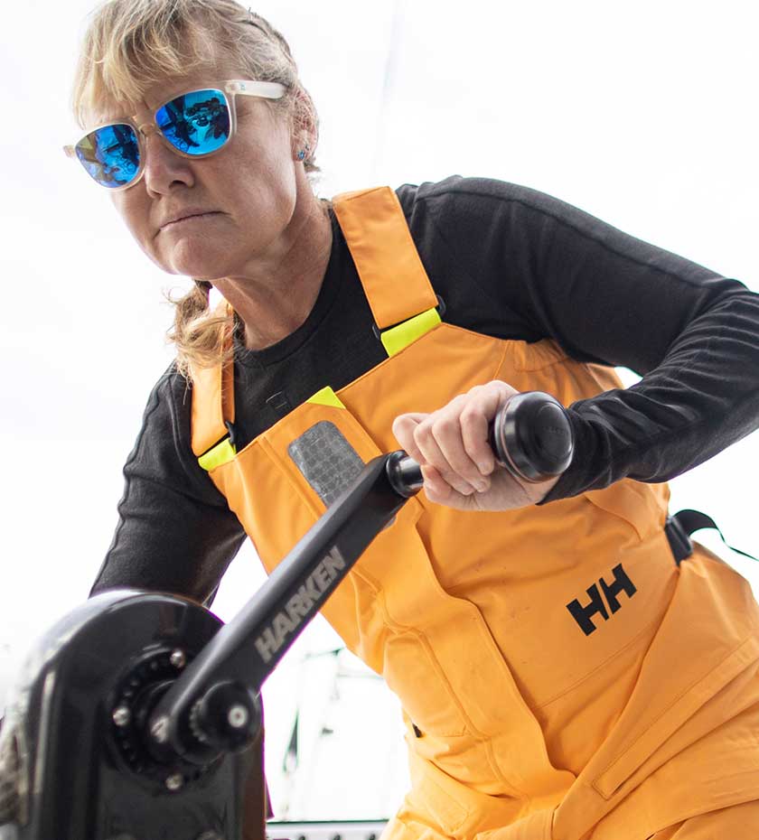 Sailing Salopettes - Female sailor in gold colour Helly Hansen sailing bib trousers winds a yacht sail winch.