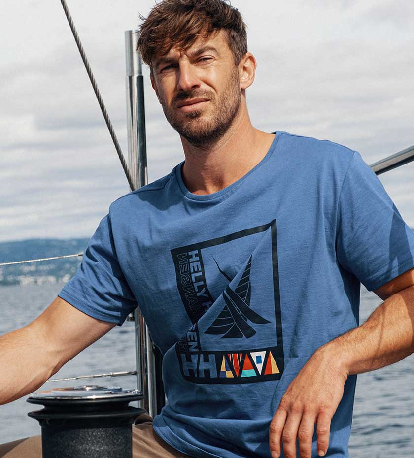 Sailinlg T- Shirts - Man wears a pale blue T-shirt with Helly hansen logo while sitting on a yacht with the sea and coastline in the background.