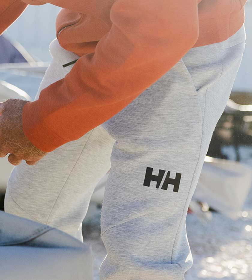 Man wears Grey HH sailing trousers with pebble beach background.