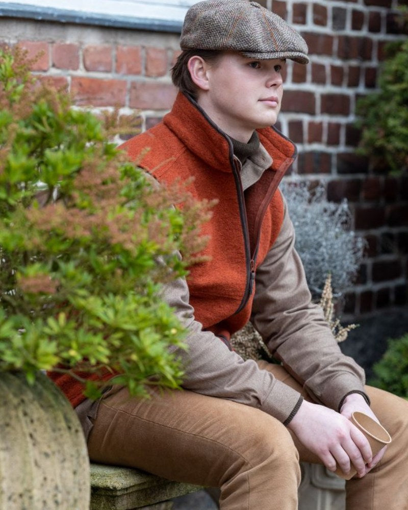 Male model wearing an Orange Laksen Gilet and tweed flat cap, sat on a bench against a brick wall.