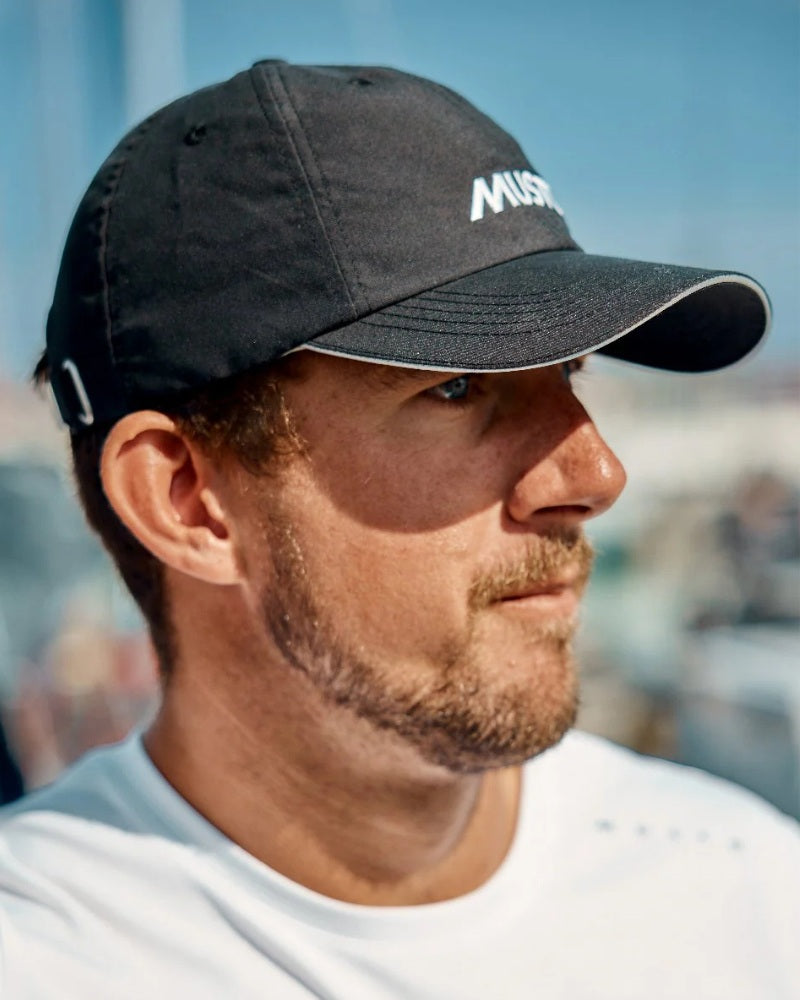 Male model wearing a black Musto branded cap with a blue sky background.