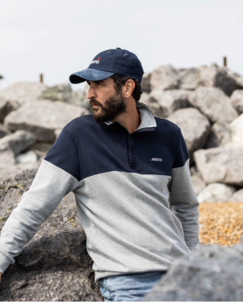 Male model leaning on a rock and looking to the left, wearing a grey and navy Musto sweatshirt and Navy Musto hat.
