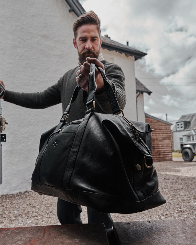 Man carrying a black Hoggs of Fife leather holdall bag outdoors in a countryside setting