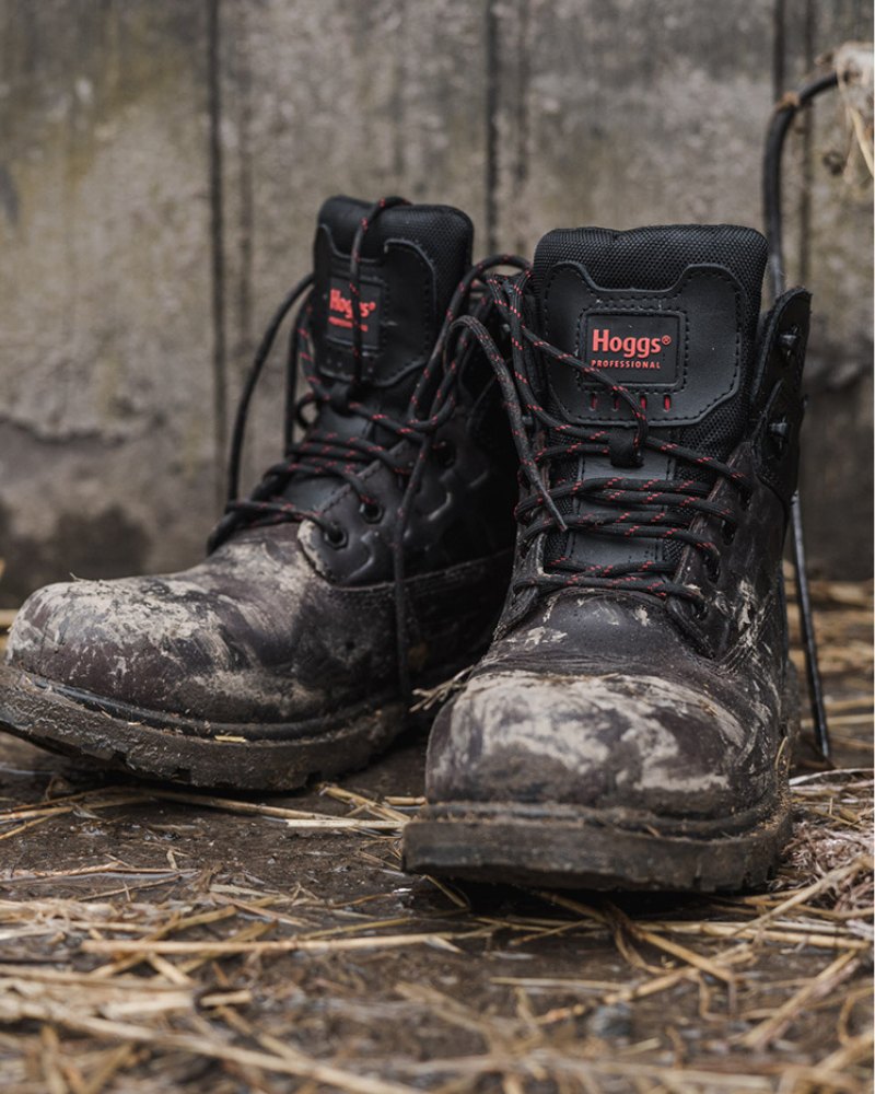 Pair of black muddy Hoggs of Fife safety boots on a mud and straw floor with a concrete background. 