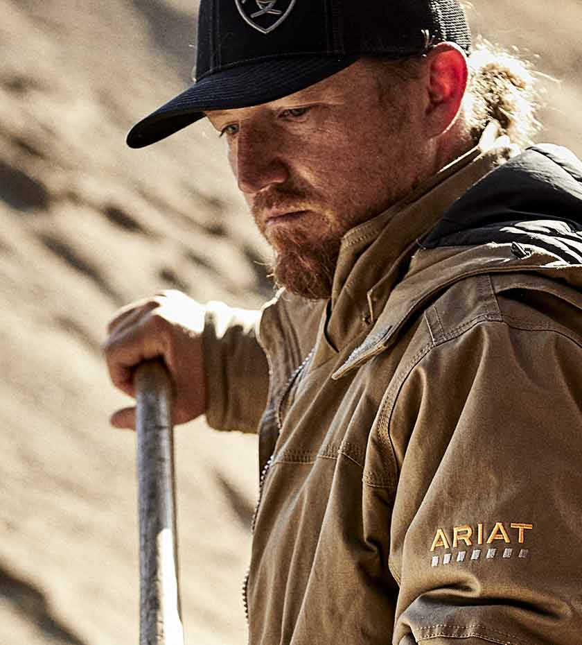 Man working with tool wears a beige hardwearing Ariat Rebar Workwear Jacket.