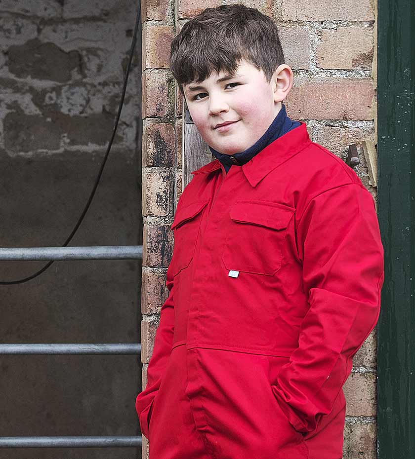 Plum boy wears red overall boilersuit whilst leaning on a brick wall.
