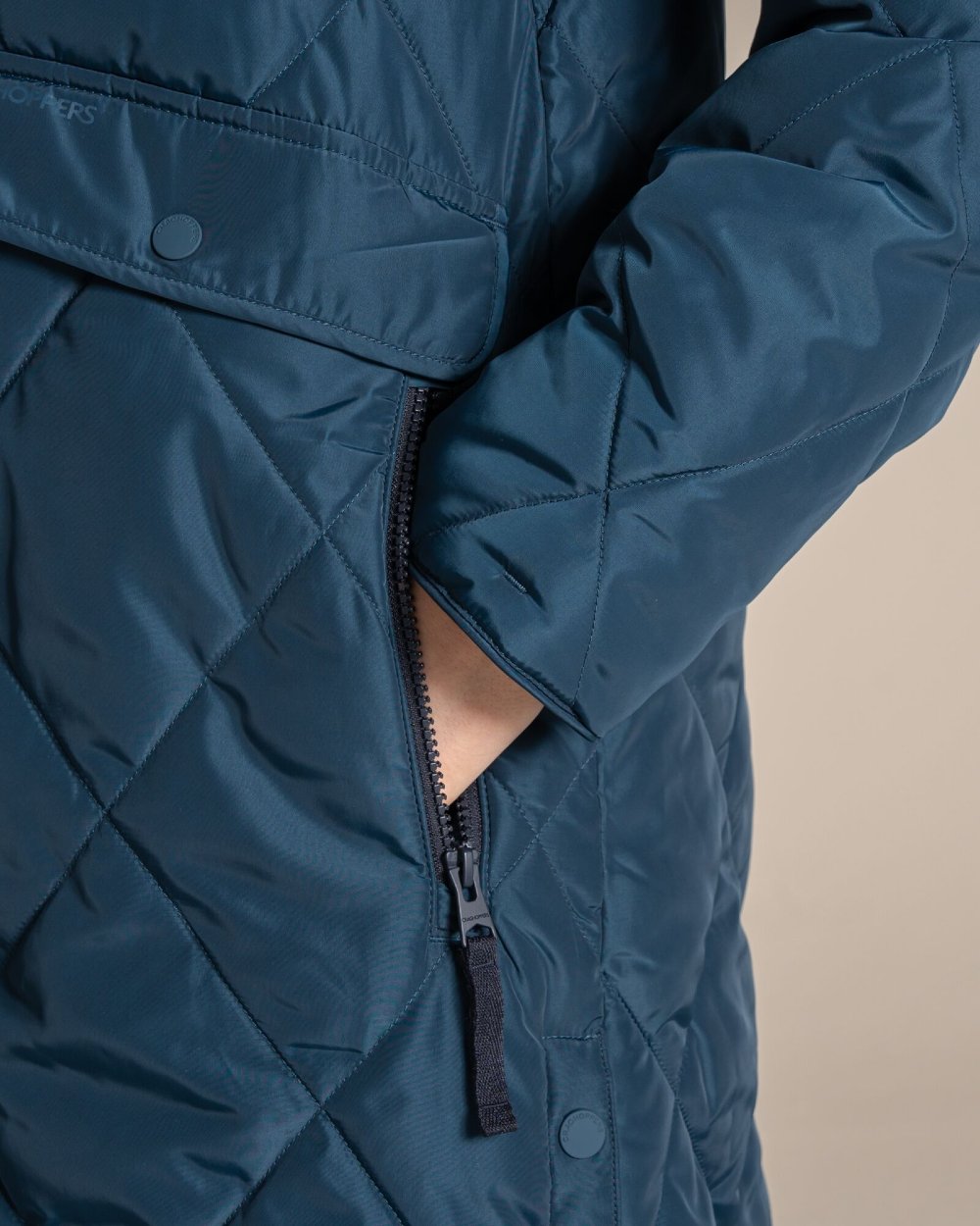Close-up of a quilted navy blue jacket with a diagonal zippered pocket and a hand tucked inside, set against a solid beige background. 