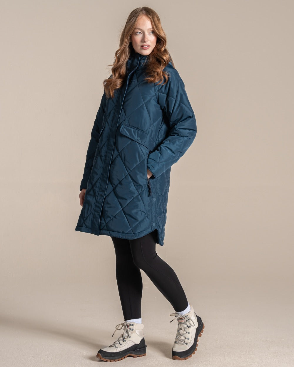 A woman modeling a mid-length quilted coat in a deep teal blue color, paired with black leggings and hiking boots, against a solid beige studio background. 