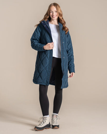 A woman modeling a mid-length quilted coat in a deep teal blue color, paired with black leggings and hiking boots, against a solid beige studio background. 