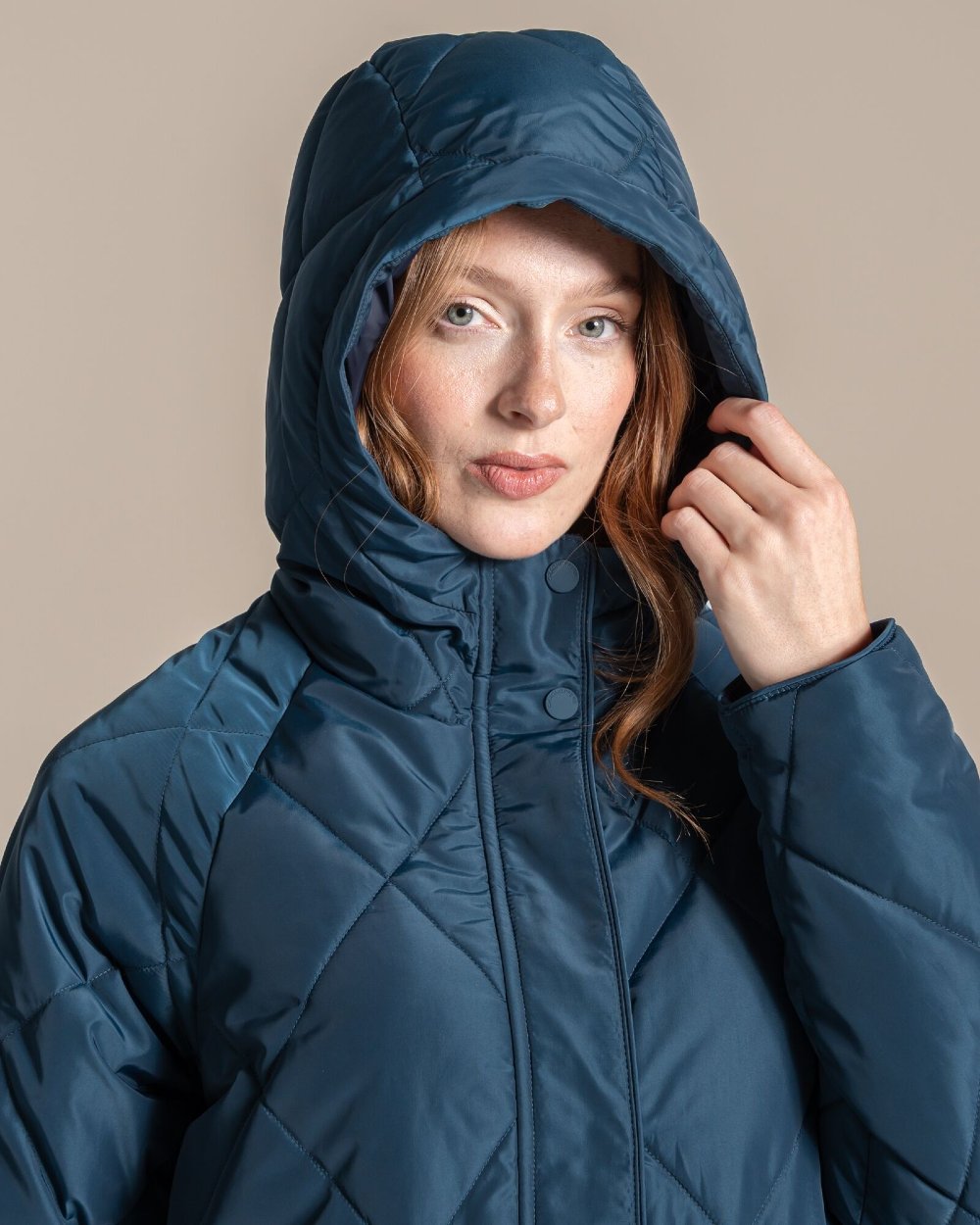 A woman wearing a navy blue hooded puffer jacket, shown from the chest up against a solid beige background. 