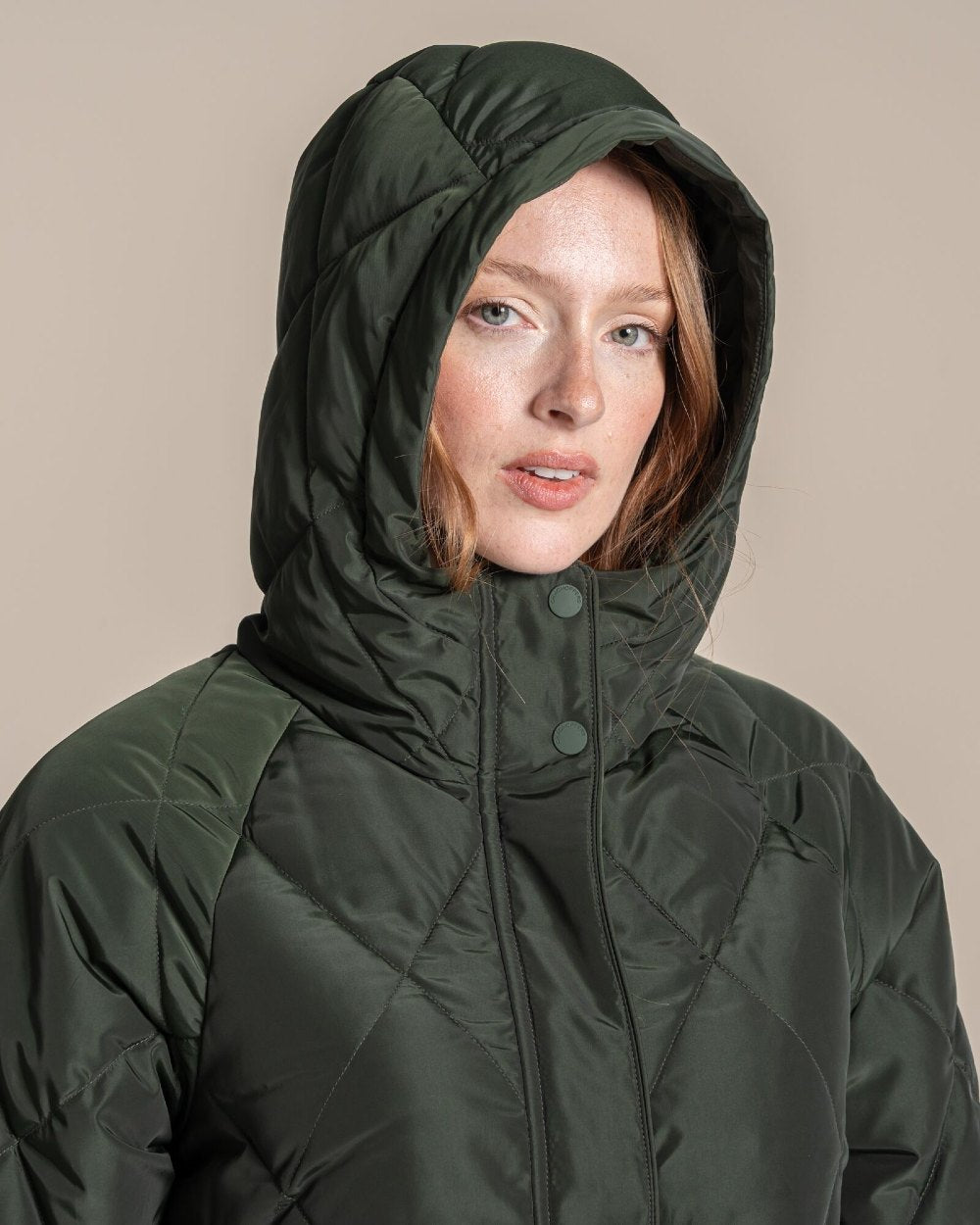 A woman modeling a dark green quilted hooded parka jacket standing against a plain beige studio background. 