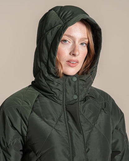 A woman modeling a dark green quilted hooded parka jacket standing against a plain beige studio background. 