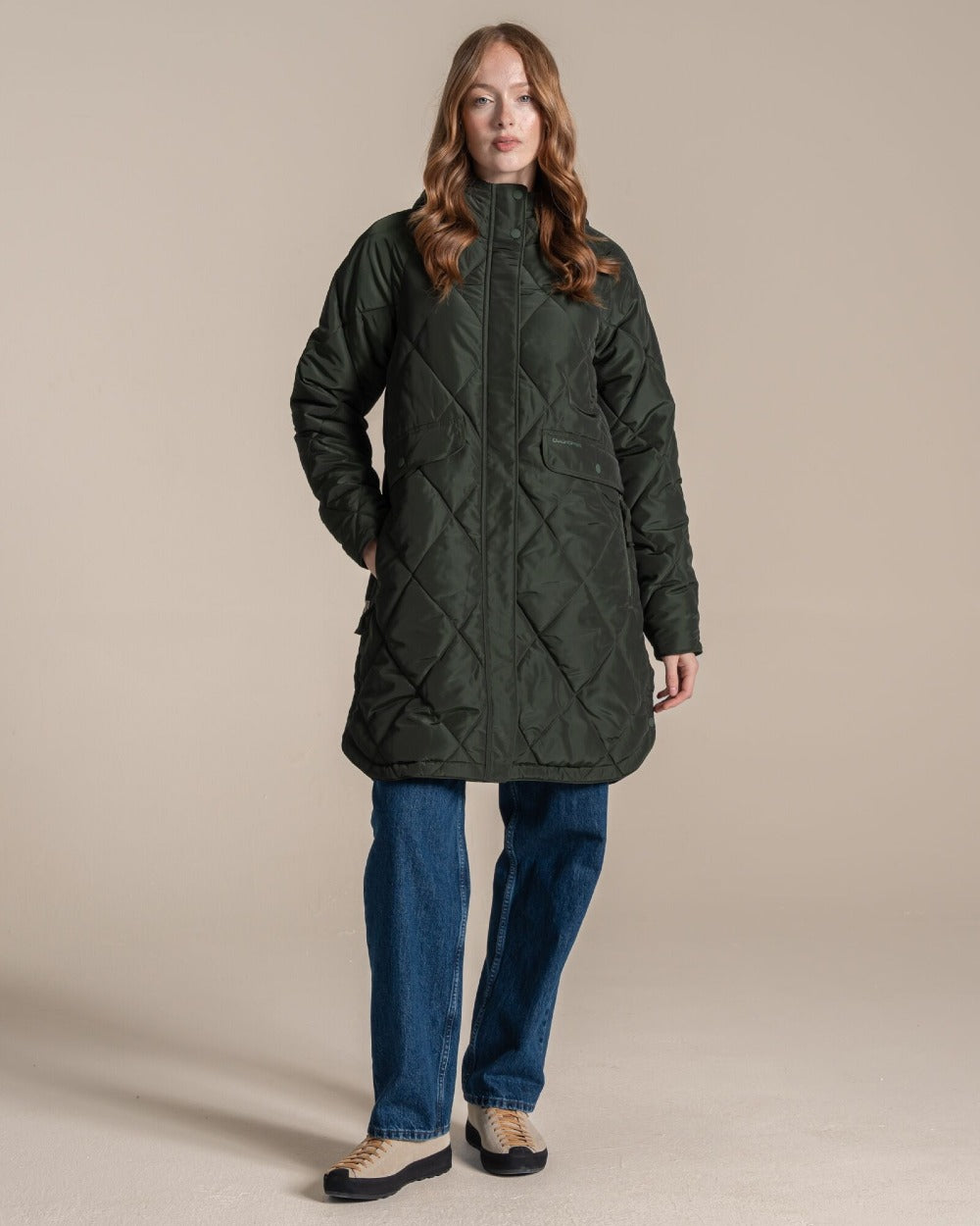 A woman modeling a dark green quilted hooded parka jacket over a beige top and blue jeans, standing against a plain beige studio background. 