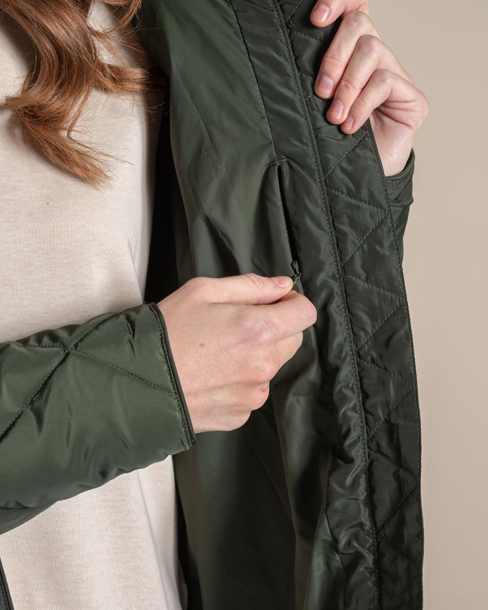 Close-up of a person wearing an olive green quilted jacket, showing the texture of the fabric and zipper detail against a neutral beige background. 