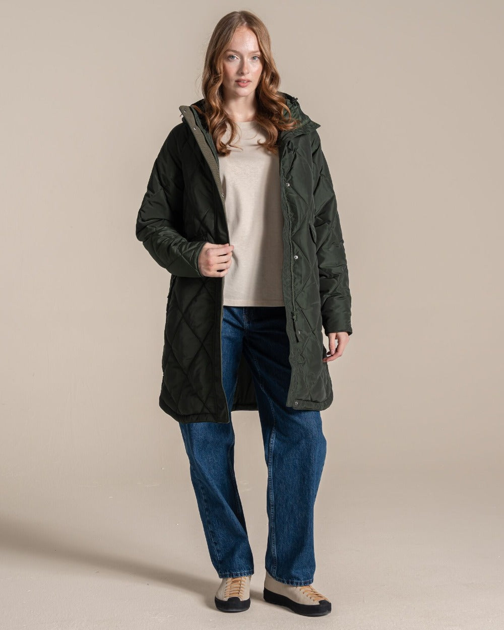 A woman modeling a dark green quilted hooded parka jacket over a beige top and blue jeans, standing against a plain beige studio background. 