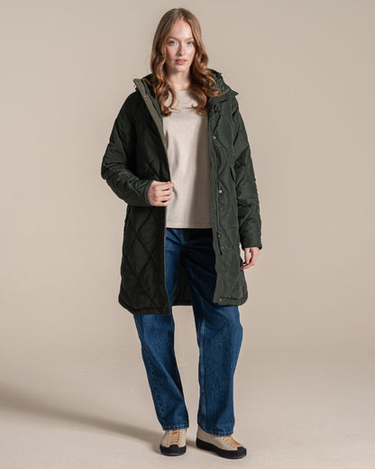 A woman modeling a dark green quilted hooded parka jacket over a beige top and blue jeans, standing against a plain beige studio background. 