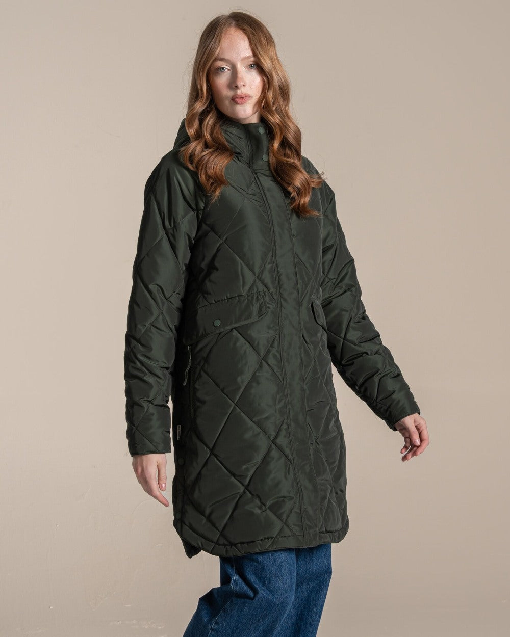 A woman modeling a dark green quilted hooded parka jacket over a beige top and blue jeans, standing against a plain beige studio background. 