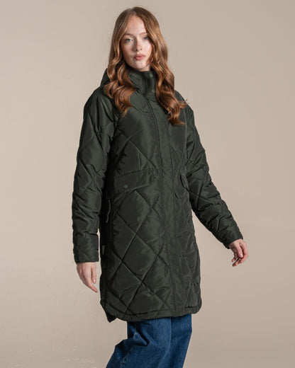 A woman modeling a dark green quilted hooded parka jacket over a beige top and blue jeans, standing against a plain beige studio background. 