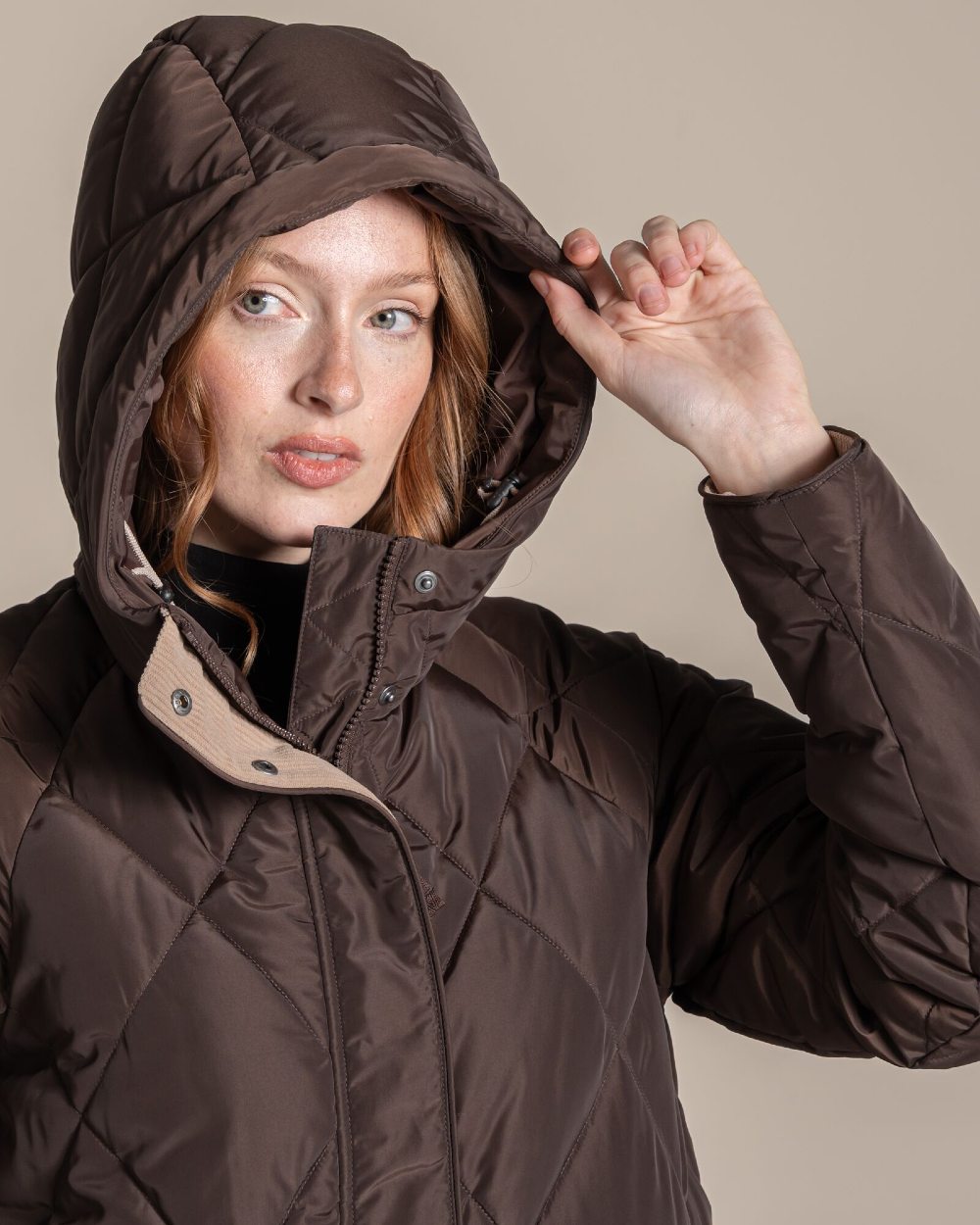 A woman modeling a dark brown diamond-quilted coat with large hood, against a solid beige studio background. 
