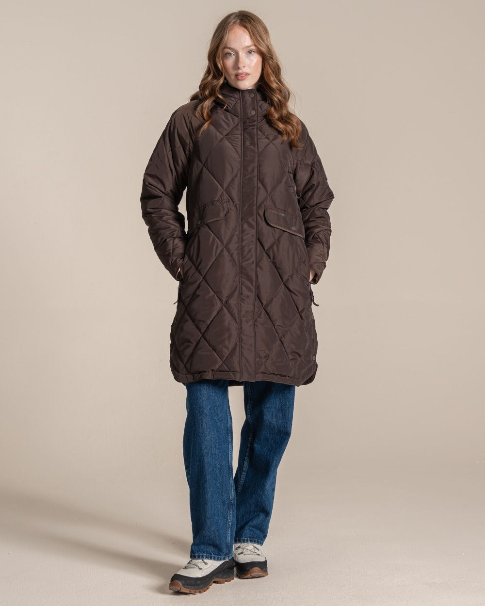 A woman modeling a dark brown diamond-quilted coat with front pockets, paired with blue jeans and white sneakers, against a solid beige studio background. 