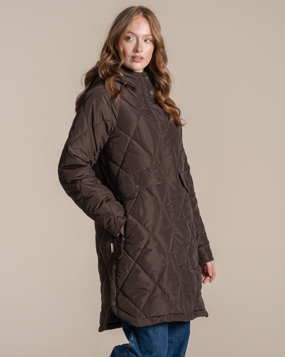 A woman modeling a dark brown diamond-quilted coat with front pockets, paired with blue jeans and white sneakers, against a solid beige studio background. 