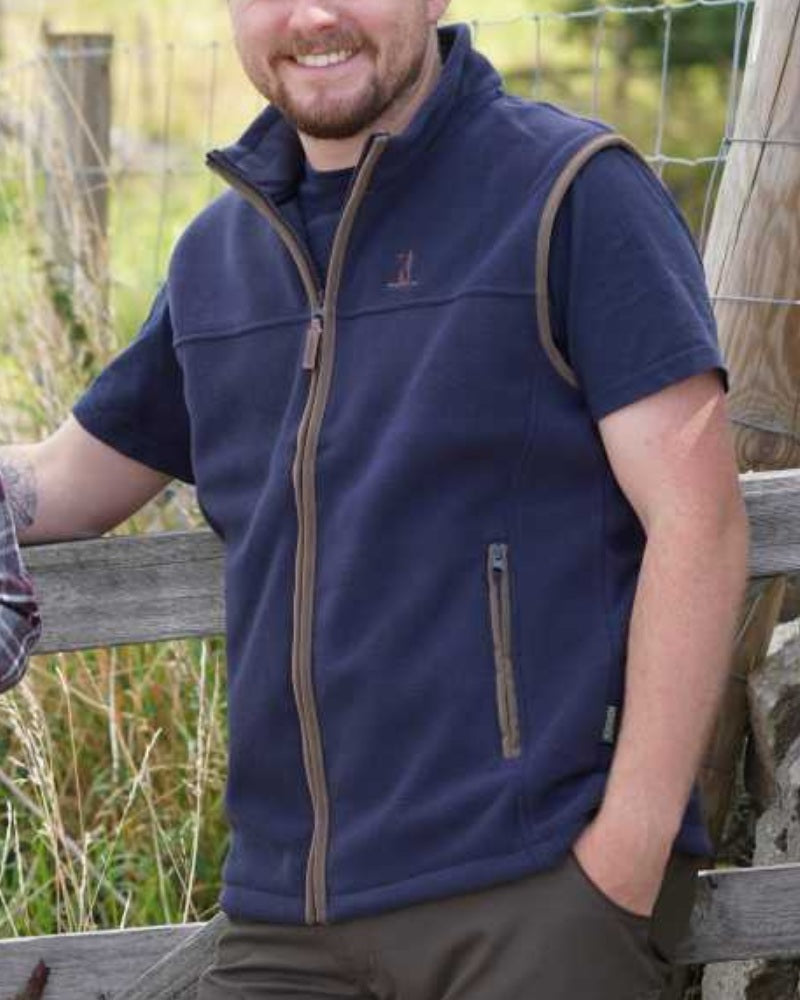 Man wearing a navy Percussion fleece gilet outdoors beside a wooden fence