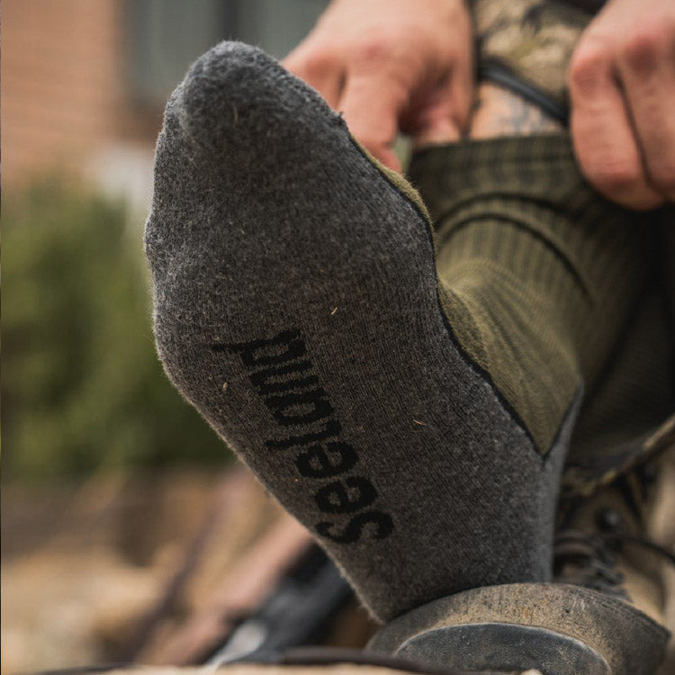A close-up of a person's foot wearing Seeland sock, with hands adjusting the sock. The Seeland brand name is clearly printed in black on the sole of the sock.
