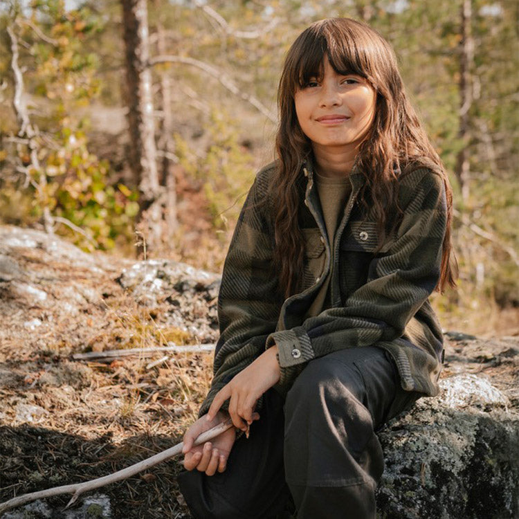 A young girl with long brown hair sits on a mossy rock in a forest, wearing a dark olive green and black plaid fleece jacket over a tan shirt and dark pants.