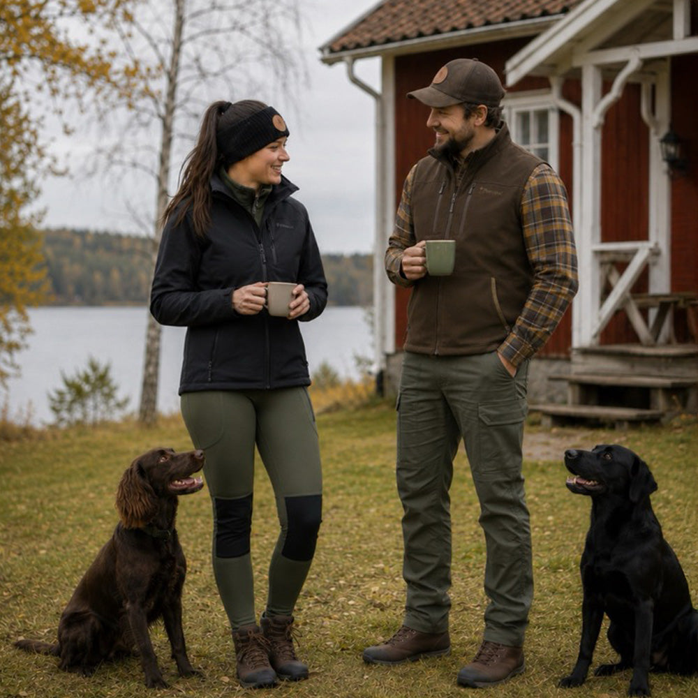 A man and a woman standing outdoors in front of a red house, each holding a mug, with two dogs sitting on the grass beside them.