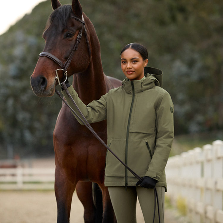 A woman in an olive green equestrian jacket and matching riding tights stands beside a brown horse in an outdoor stable setting, holding the horse's reins.